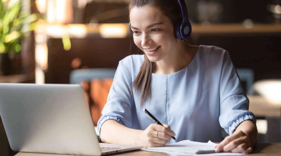 smiling woman wearing headphones taking notes while attending an online meeting - learning management system​